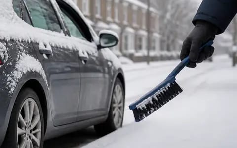 A gloved hand holds a blue snow brush beside a grey car still covered in snow