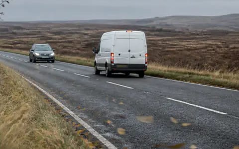A white high-roof van and a grey car travel along a wet, open moorland road in grey, windy weather