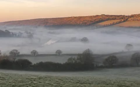 Mist settles over a chilled valley floor where cold air lies among hedgerows and trees