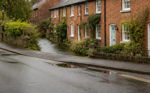 A stream running beside a wet road past red-brick cottages, the nearby water that flood risk depends on