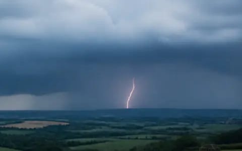 Distant forked lightning flashing beneath a dark storm cloud above rolling green countryside