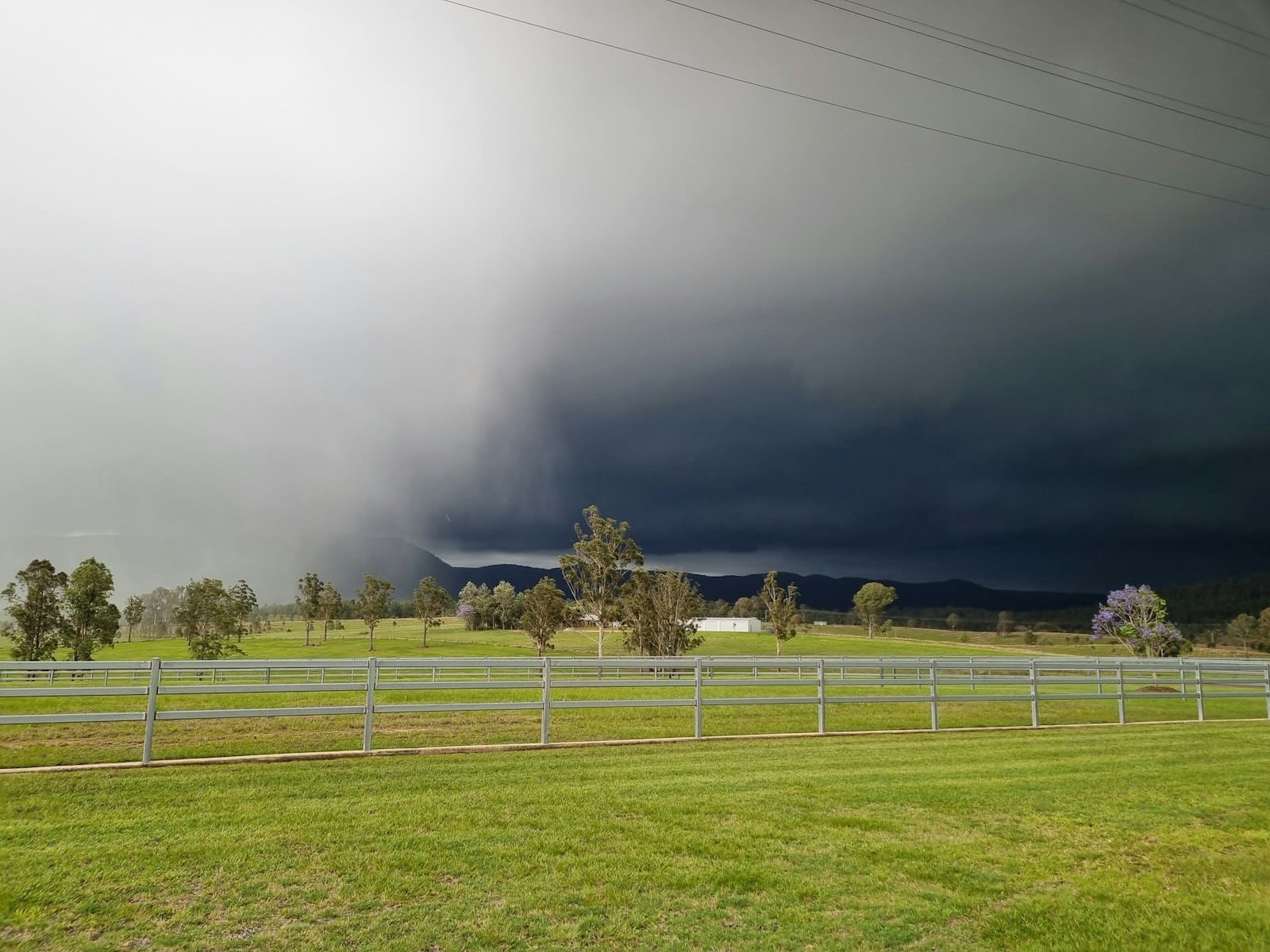 A dark wall of rain cloud moving across open grassland and a white-railed paddock, with lighter sky to the left