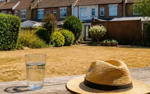 A glass of water and a straw hat on a sunny table beside a scorched lawn