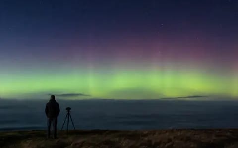 A photographer with a tripod watches green and pink aurora glowing above the dark sea