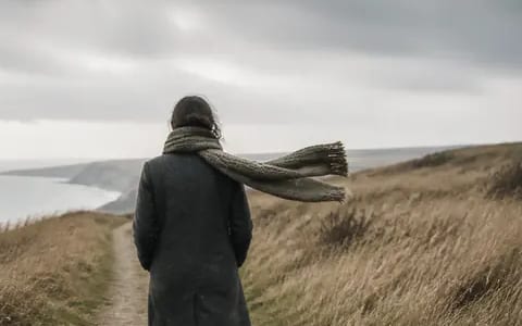 A long knitted scarf streams sideways in a strong coastal wind on a grey, exposed clifftop